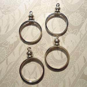 Könnte beinhalten: Vier silberfarbene Münzrahmen-Anhänger mit dekorativem Aufsatz und kleiner Schlaufe zum Befestigen an einer Kette. Die kreisförmigen Rahmen haben einen strukturierten Rand. Die Anhänger werden auf einem gemusterten Hintergrund angezeigt.