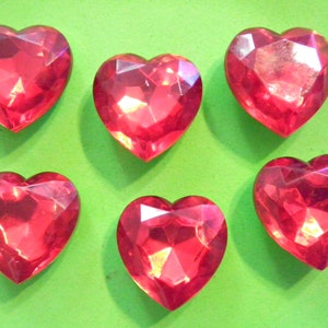 May include: Six faceted, heart-shaped, red gemstones are arranged on a bright green background. The gems have a sparkling, jewel-like appearance, catching the light with their multiple facets. The hearts are evenly spaced, creating a visually appealing pattern.