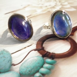 May include: Two silver rings with large, oval, iridescent blue-purple stones. The rings are set against a backdrop of a wooden ring and turquoise beads, creating a contrast of textures and colors. The stones have a glossy finish.