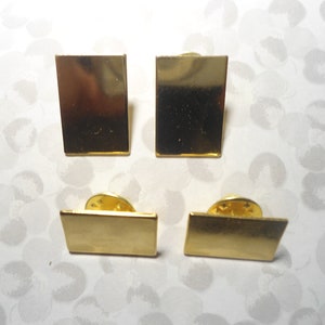 May include: Four gold-colored rectangular lapel pins. Two are upright, and two are horizontal. Each pin has a gold-colored pin back. The pins are on a white background with a gray floral pattern.