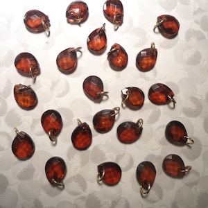 May include: A collection of faceted, teardrop-shaped amber-colored beads with gold-colored loops. The beads are scattered across a patterned, off-white surface, showcasing their glossy, transparent appearance. These beads are suitable for jewelry making.