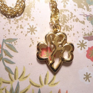 May include: A gold-coloured necklace with a pendant depicting a stylized figure within a clover-shaped frame. The chain has a twisted rope design. The background features a floral pattern in pink, green, and gold.