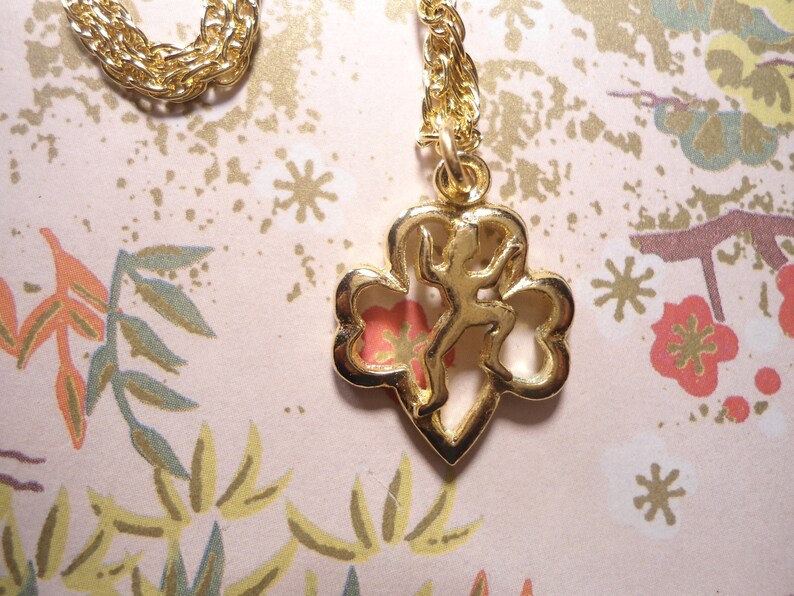 May include: A gold-coloured necklace with a pendant depicting a stylized figure within a clover-shaped frame. The chain has a twisted rope design. The background features a floral pattern in pink, green, and gold.