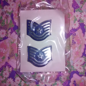 Peut inclure: Deux insignes de grade militaire bleus et argentés, chacun avec une étoile dans un motif de crête, sont présentés sur une carte blanche, scellés dans un sac en plastique transparent. Le fond est un tissu à motifs floraux.