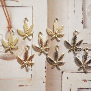 Könnte beinhalten: Goldfarbene Metallanhänger in Form von Cannabisblättern mit einer Schlaufe zur Schmuckherstellung. Das Bild zeigt auch eine Halskette mit einem weißen Anhänger.