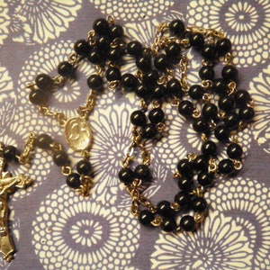 May include: A rosary with black beads and a silver crucifix. The rosary features a silver centerpiece with an image and is displayed on a patterned background.