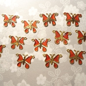 May include: A collection of orange and gold butterfly charms. The butterfly charms have a gold outline and orange enamel wings. The charms are scattered on a light gray background with a floral pattern.