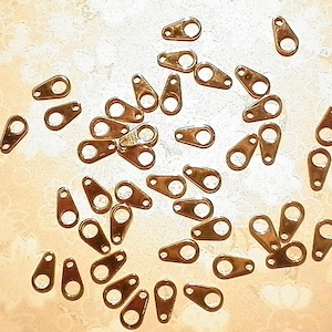 Puede incluir: Un conjunto de pequeños herrajes metálicos de color cobre, cada uno con un extremo redondeado y un agujero para fijar. Estos herrajes están dispersos sobre una superficie clara, ideales para la creación de joyas o proyectos de artesanía.