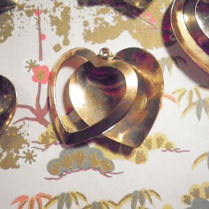 May include: Gold-toned heart-shaped pendants. The pendants feature a layered design with an inner heart shape visible within the outer heart. The background is a patterned paper with floral and cloud motifs.