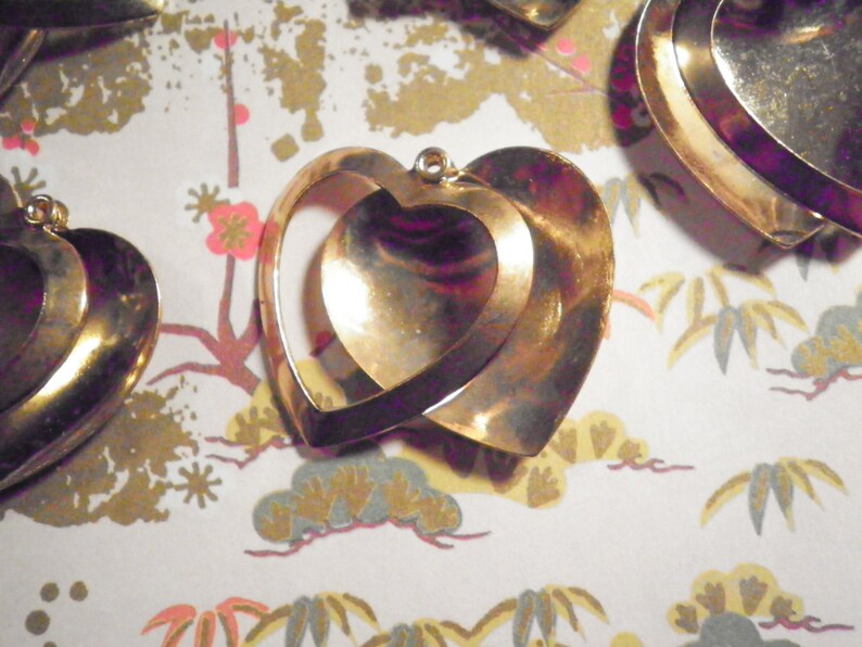 May include: Gold-toned heart-shaped pendants. The pendants feature a layered design with an inner heart shape visible within the outer heart. The background is a patterned paper with floral and cloud motifs.