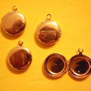 May include: Four gold-colored, round lockets with a loop at the top for a chain. One locket is open, revealing a mirrored interior. One locket has the text "© WALT DISNEY PRODUCTIONS" on the back. The lockets are on a yellow background.