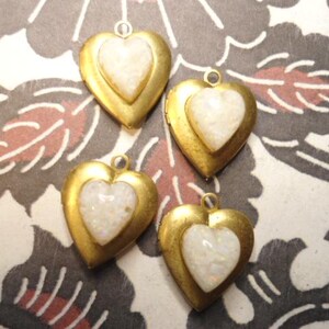 May include: Four heart-shaped pendants with a gold-colored exterior and a white, textured heart-shaped inlay. Each pendant has a small loop at the top for attaching to a chain or other jewelry. The pendants are displayed on a patterned background.