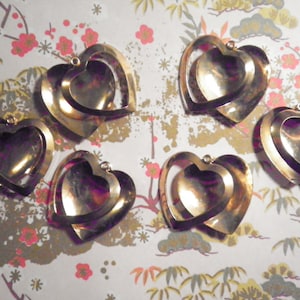 May include: Six gold-colored heart-shaped pendants with a layered design. Each heart has a small loop at the top for hanging. The pendants are arranged on a floral patterned background with pink and gold accents.