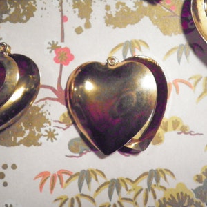 May include: Gold-colored heart-shaped lockets with a hinged design, displayed on a patterned background. The lockets have a shiny, reflective surface and a small loop at the top for a chain. The background features a floral and foliage pattern.
