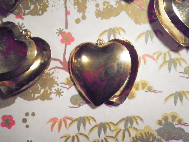 May include: Gold-colored heart-shaped lockets with a hinged design, displayed on a patterned background. The lockets have a shiny, reflective surface and a small loop at the top for a chain. The background features a floral and foliage pattern.