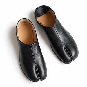Könnte beinhalten: Ein Paar schwarze Slipper aus Leder mit einem markanten Zehenschlitz-Design. Die Schuhe haben eine glatte, dunkle Außenfläche mit einem beigefarbenen Innenfutter und eine flache Silhouette, die eine klare und einfache Konstruktion aufweist.