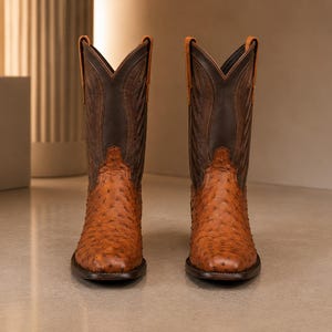 Könnte beinhalten: Ein Paar wadenhohe Cowboystiefel mit einem strukturierten, hellbraunen Straußenlederfuß mit markantem Federkielmuster und einem dunkelbraunen Lederschaft. Die Stiefel haben eine quadratische Zehenpartie, Zuglaschen und aufwendige Nähte am Schaft.