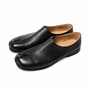 May include: A pair of black leather slip-on shoes featuring a distinct split-toe design. The shoes have a smooth, matte black finish, a low-profile silhouette, and a tan interior lining. The construction includes a central seam running from the split toe.