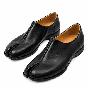 May include: A pair of black leather slip-on shoes featuring a distinct split-toe design. The shoes have a smooth, dark exterior finish, a low heel, and a tan interior lining. The construction includes a central seam running from the toe to the opening.