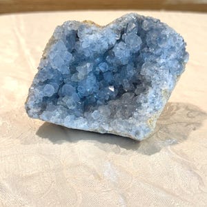 May include: A celestite geode featuring a cluster of pale blue, translucent, angular crystals forming a hollow cavity. The exterior of the mineral specimen is a rough, light tan, rocky matrix that supports the crystalline structure.