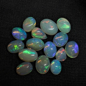 May include: A collection of thirteen oval and pear-shaped cabochon opals. Each gemstone features a translucent white base with vibrant flashes of iridescent color, including green, blue, and orange, visible across the smooth, polished surfaces.