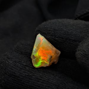 May include: A raw, uncut opal gemstone featuring a translucent body with vibrant flashes of neon orange, bright green, and yellow play-of-color. The stone has an irregular, angular shape with natural, unpolished edges and a textured, rocky surface.
