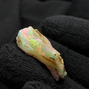 May include: A raw, uncut opal gemstone featuring a translucent, milky white base with vibrant flashes of green, orange, and pink play-of-color. The stone has an irregular, jagged shape with natural surface textures and small dark inclusions.