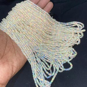 Könnte beinhalten: Mehrere Stränge aus kleinen, runden, durchscheinenden Opalperlen mit einer milchig-weißen Grundfarbe und irisierenden Lichtreflexen in Rosa, Grün und Blau. Die Perlen sind in langen, gleichmäßigen Reihen aufgereiht und wirken schimmernd.