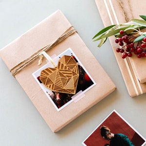 May include: Two brown gift boxes wrapped with twine and a sprig of greenery with red berries. One box has a wooden heart ornament with a geometric pattern and a photo attached. A white gift tag with the text "To The Best Dad!" is being written on with a gold pen.