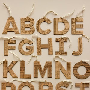 Puede incluir: Adornos de madera en forma de alfabeto con un patrón geométrico. Cada letra es un adorno separado con una cuerda para colgar.