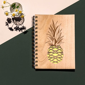 May include: A wooden spiral bound notebook with a laser-cut pineapple design. The pineapple is yellow and the notebook is brown.