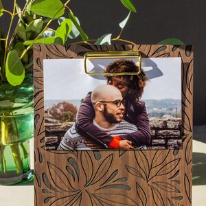 May include: A wooden photo frame with a floral design and a gold metal clip. The frame holds a photo of a couple embracing.