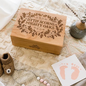 Puede incluir: Una caja de madera con un acabado marrón claro y un diseño floral grabado en la tapa. La tapa dice "From Little Acorns Mighty Oaks Grow". La caja está sobre una superficie con estampado beige y blanco con un par de botines de bebé de punto marrón, una manta blanca y un pequeño jarrón con una rama de vegetación.
