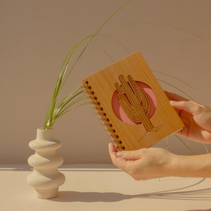 Puede incluir: Un cuaderno de espiral de madera con un diseño de cactus cortado con láser en un círculo rosa. El cuaderno está siendo sostenido por manos.