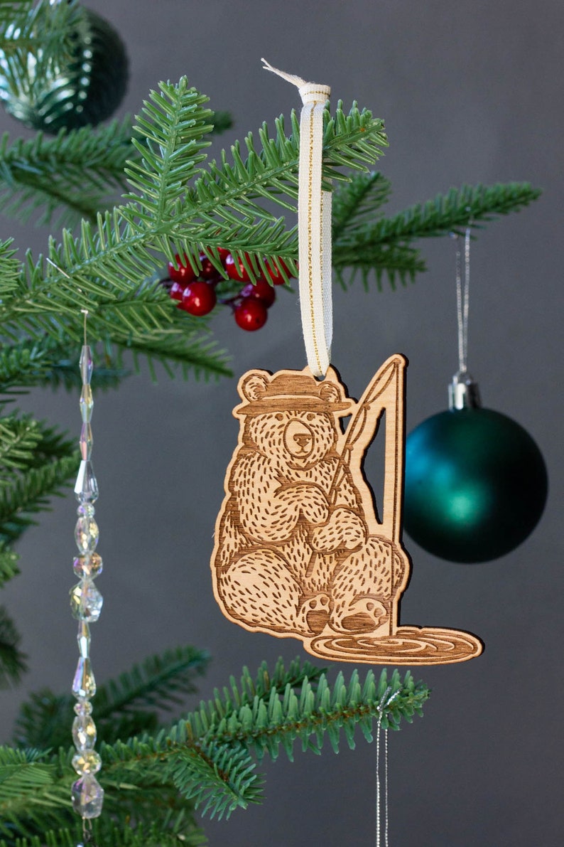Pode incluir: Adorno de Natal de madeira com um urso usando um chap&eacute;u e segurando uma vara de pesca. O urso est&aacute; sentado em uma po&ccedil;a de &aacute;gua com ondas. O adorno est&aacute; pendurado em uma fita branca em uma &aacute;rvore de Natal verde.