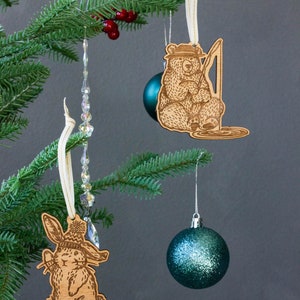 Pode incluir: Tr&ecirc;s enfeites de Natal pendurados em uma &aacute;rvore de Natal artificial verde. Os enfeites s&atilde;o um urso de madeira com uma vara de pesca, um coelho de madeira com um chap&eacute;u e uma bola de cristal azul esverdeada com glitter.