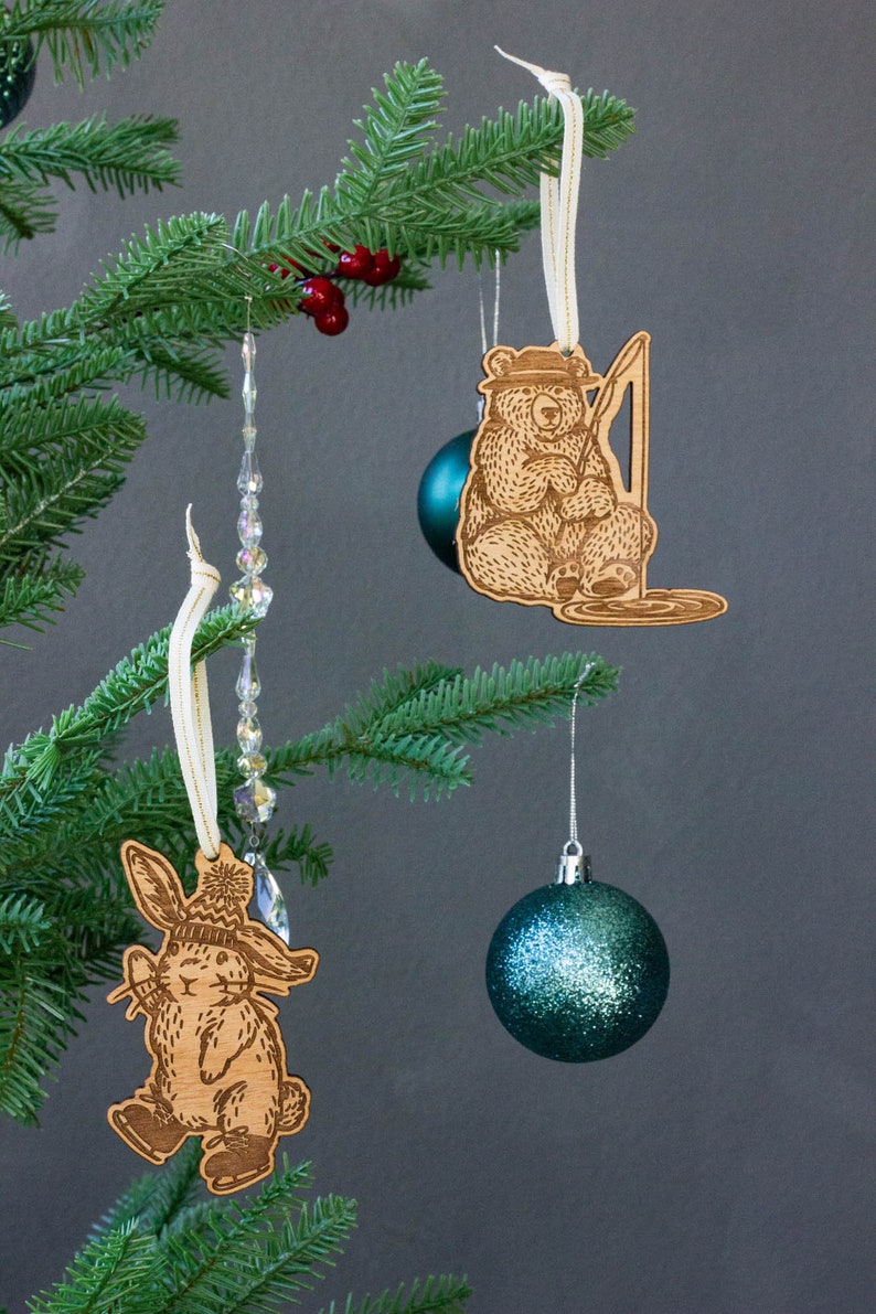 Pode incluir: Tr&ecirc;s enfeites de Natal pendurados em uma &aacute;rvore de Natal artificial verde. Os enfeites s&atilde;o um urso de madeira com uma vara de pesca, um coelho de madeira com um chap&eacute;u e uma bola de cristal azul esverdeada com glitter.