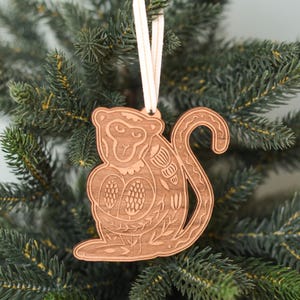 May include: Wooden ornament of a monkey with intricate floral details, hanging from a white string on a green Christmas tree.