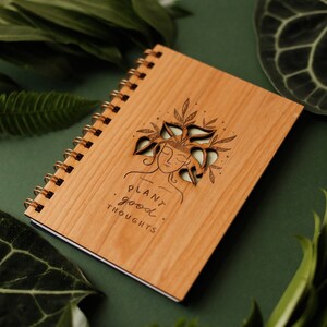 May include: A wooden spiral-bound notebook with a laser-cut design of a woman's face with plants growing from her head. The notebook has the words "PLANT good THOUGHTS" printed on it. The background is a dark green.