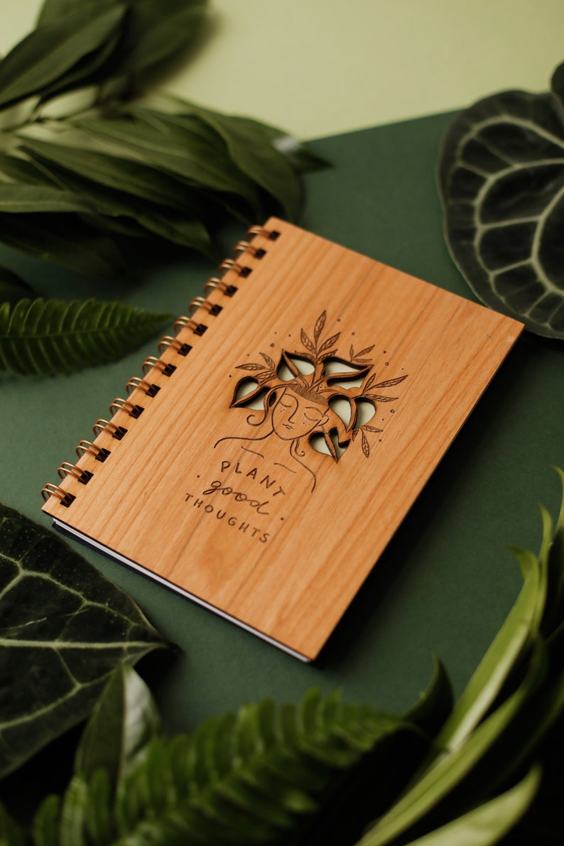 May include: A wooden spiral-bound notebook with a laser-cut design of a woman's face with plants growing from her head. The notebook has the words "PLANT good THOUGHTS" printed on it. The background is a dark green.