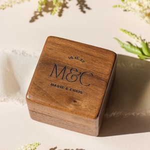 Puede incluir: Una caja de anillos cuadrada de madera con texto grabado. La caja es de color marrón cálido y presenta las iniciales "M&C" con la fecha "10.15.22" arriba y los nombres "Marie & Chris" abajo. El fondo incluye flores blancas y sombras suaves.