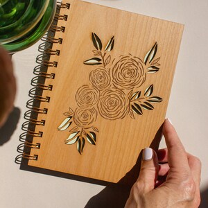 Puede incluir: Un diario de madera con encuadernaci&oacute;n en espiral y un dise&ntilde;o floral cortado con l&aacute;ser. El dise&ntilde;o presenta un grupo de rosas y hojas de color marr&oacute;n claro.