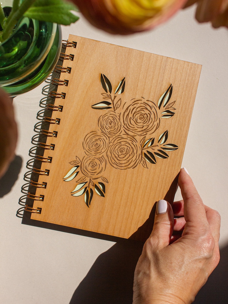 Puede incluir: Un diario de madera con encuadernaci&oacute;n en espiral y un dise&ntilde;o floral cortado con l&aacute;ser. El dise&ntilde;o presenta un grupo de rosas y hojas de color marr&oacute;n claro.