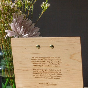 May include: A wooden plaque with a heartfelt message engraved in a light brown font. The message expresses love and appreciation for someone special. The plaque is propped up on a gold metal stand. The background features a bouquet of white flowers in a green vase.