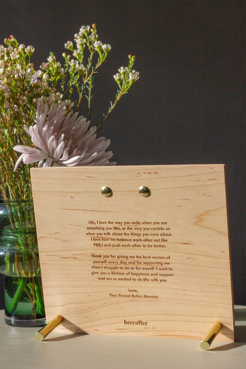 May include: A wooden plaque with a heartfelt message engraved in a light brown font. The message expresses love and appreciation for someone special. The plaque is propped up on a gold metal stand. The background features a bouquet of white flowers in a green vase.