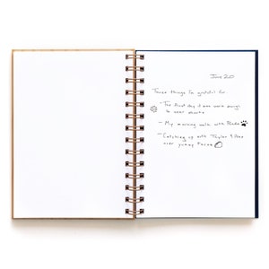 May include: An open notebook with a blue cover and a spiral binding. The right page has handwritten text that reads "June 20. Three things I'm grateful for: -The first day it was warm enough to wear shorts -My morning walk with Panda -Catching up with Taylor & Alex over yummy tacos".