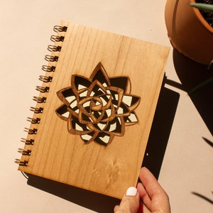 Puede incluir: Un cuaderno de espiral de madera con un dise&ntilde;o de suculenta cortado con l&aacute;ser. La cubierta marr&oacute;n claro presenta una suculenta detallada y en capas en tonos marr&oacute;n, negro y crema. El cuaderno es sostenido por una persona.