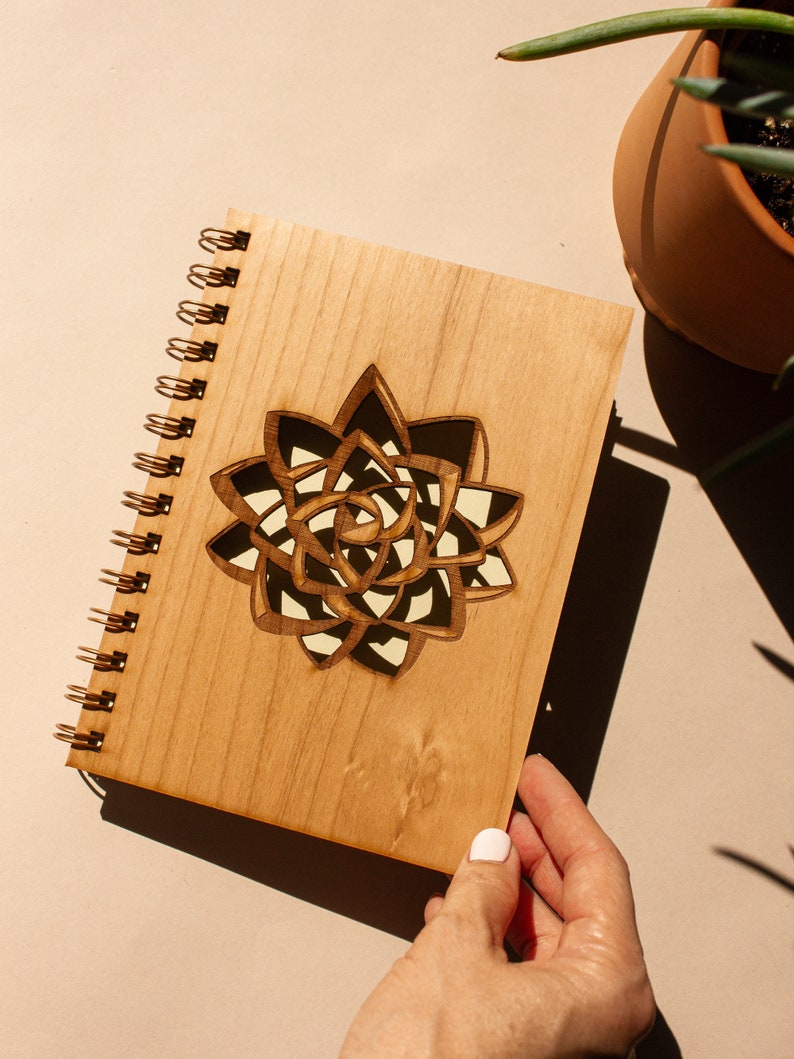 Puede incluir: Un cuaderno de espiral de madera con un dise&ntilde;o de suculenta cortado con l&aacute;ser. La cubierta marr&oacute;n claro presenta una suculenta detallada y en capas en tonos marr&oacute;n, negro y crema. El cuaderno es sostenido por una persona.