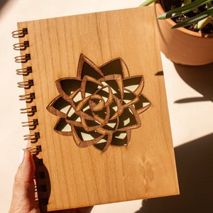 Puede incluir: Un cuaderno de espiral de madera con un dise&ntilde;o de suculenta cortado con l&aacute;ser. El cuaderno tiene una cubierta marr&oacute;n claro y una encuadernaci&oacute;n en espiral dorada. El dise&ntilde;o de la suculenta es marr&oacute;n oscuro con un interior verde claro.