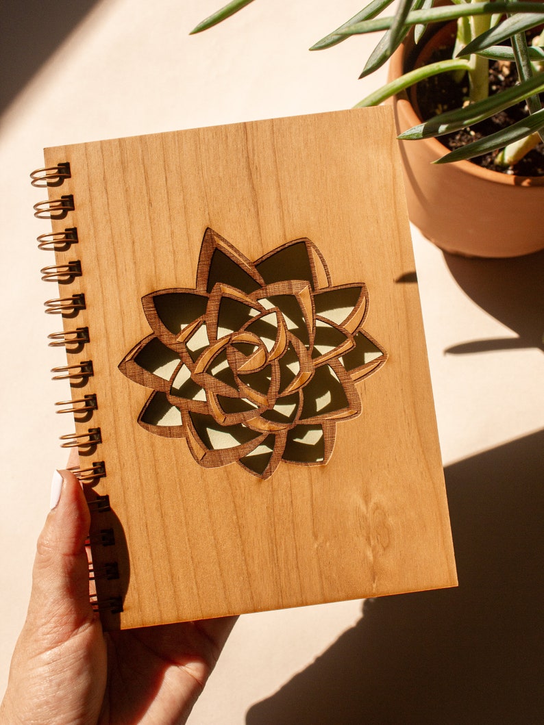 Puede incluir: Un cuaderno de espiral de madera con un dise&ntilde;o de suculenta cortado con l&aacute;ser. El cuaderno tiene una cubierta marr&oacute;n claro y una encuadernaci&oacute;n en espiral dorada. El dise&ntilde;o de la suculenta es marr&oacute;n oscuro con un interior verde claro.